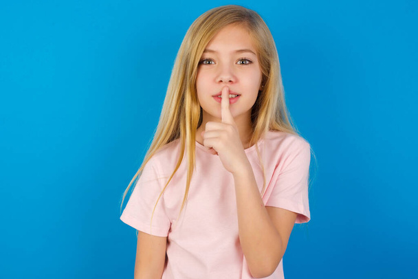Garota branca surpreso vestindo camisa rosa contra a parede azul faz gesto de silêncio, mantém o dedo sobre os lábios e parece misterioso para a câmera - Foto, Imagem