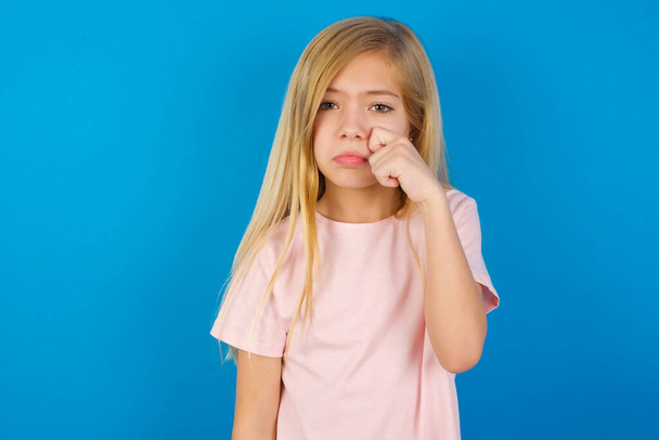 Déçu enfant caucasien déçu portant chemise rose contre les larmes lingettes murales bleues se tient stressé avec une expression sombre. Émotion négative - Photo, image