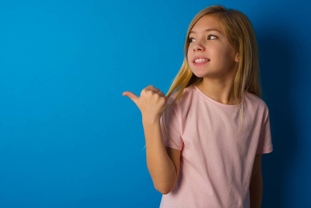 Caucásico niño niña usando rosa camisa contra azul de la pared puntos de distancia y da consejos demuestra publicidad - Foto, imagen