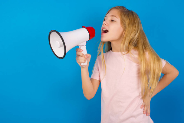 Funny Caucasian kid girl wearing pink shirt against blue wall People sincere emotions lifestyle concept. Mock up copy space. Screaming in megaphone. - Foto, Imagem