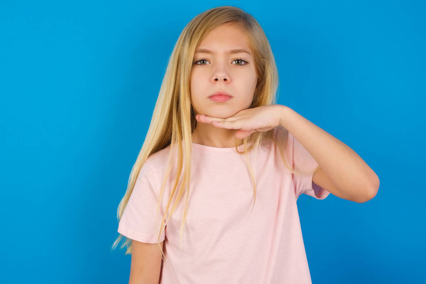 blank kind meisje dragen roze shirt tegen blauwe muur snijden keel met de hand als mes, dreigen agressie met woedend geweld. - Foto, afbeelding