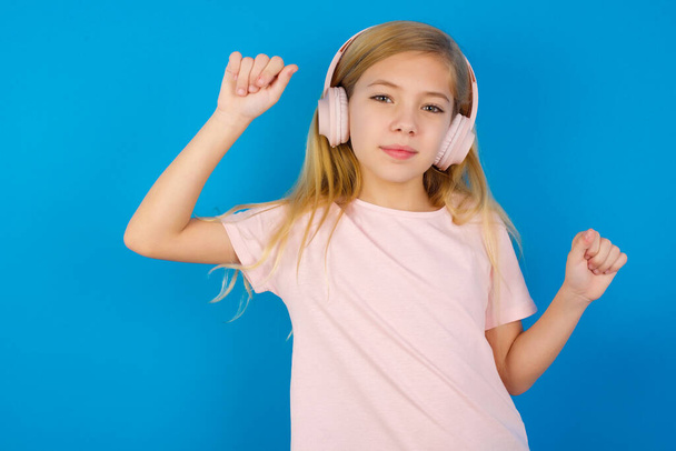 Carefree Καυκάσιος κορίτσι φορώντας φόρεμα κατά μπλε τοίχο παχιά γενειάδα και toothy χαμόγελο αυξάνει τους χορούς αγκαλιά ανέμελες κινήσεις με ρυθμό της μουσικής ακούει μουσική από playlist μέσω ακουστικών - Φωτογραφία, εικόνα