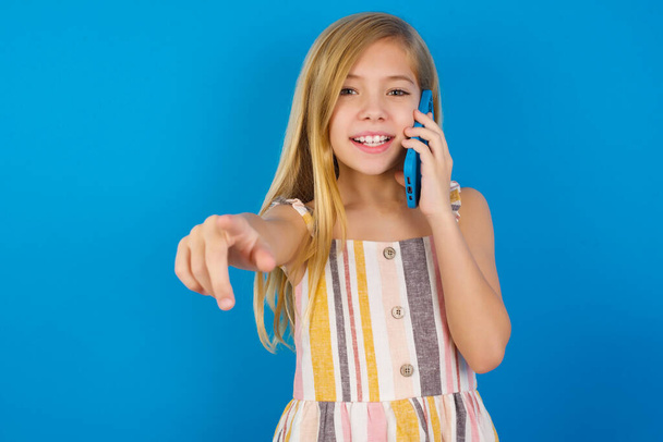 Позитивная кавказская девочка в платье на синей стене указывает прямо перед камерой имеет телефонный разговор улыбается широко любит говорить долгие часы. Ты присоединишься ко мне - Фото, изображение