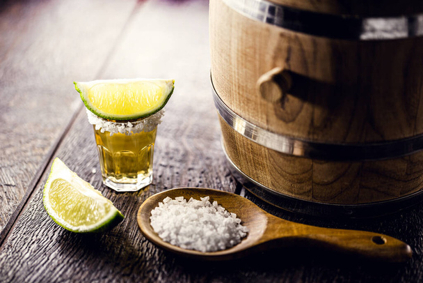 стакан текилы, дистиллированный напиток с солью и лимоном, рядом дубовый бочонок - Фото, изображение