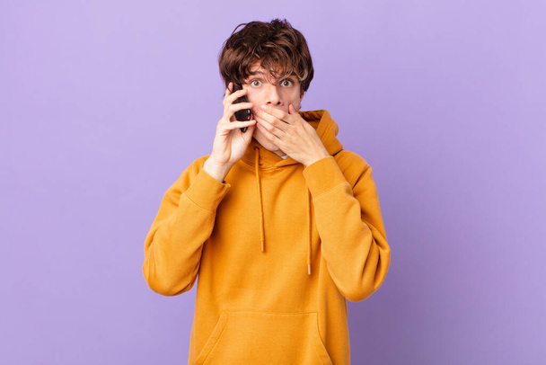 young man holding a cell covering mouth with hands with a shocked - Foto, afbeelding