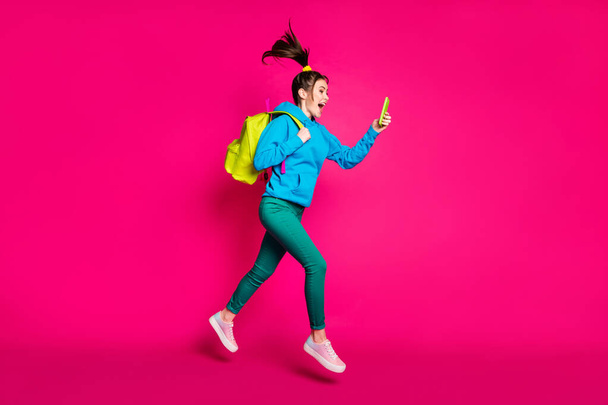 Full length photo of excited funky young lady wear blue sweater backpack looking modern device isolated pink color background - Foto, Imagem