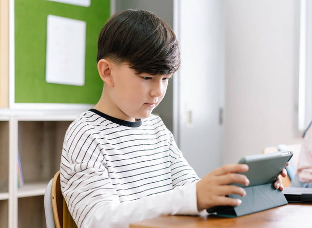İlkokulda sınıf arkadaşlarıyla masada otururken dijital tabletle konsantre olan küçük bir çocuğun portresi. Eğitim, Teknoloji ve İnternet kavramı - Fotoğraf, Görsel