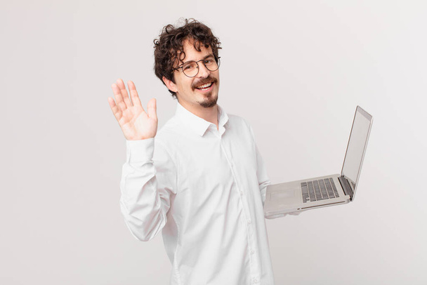 young man with a laptop smiling happily, waving hand, welcoming and greeting you - Foto, afbeelding