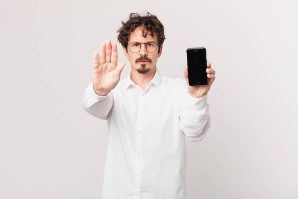 young businessman with a cell looking serious showing open palm making stop gesture - Photo, image