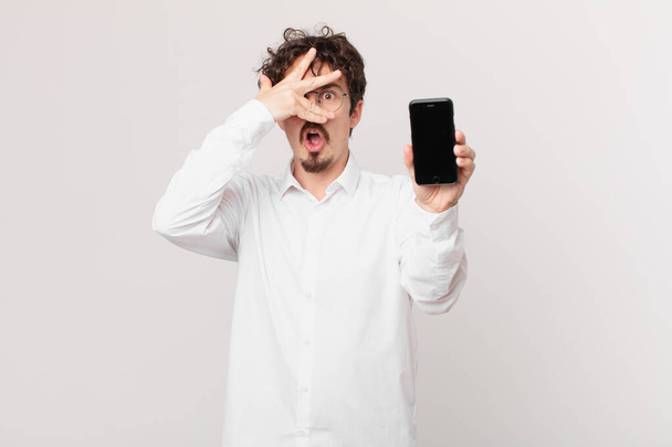 young businessman with a cell looking shocked, scared or terrified, covering face with hand - 写真・画像