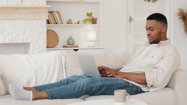 Estudiante africano enfocado tumbado en el sofá con computadora portátil escribiendo navegación en el estudio web de forma remota desde casa en cuarentena. Afro hombre negro freelancer trabajando con el ordenador sentado en el sofá de compras en línea - Metraje, vídeo