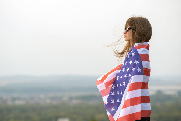 Young happy american woman with long hair holding waving on wind USA national flag on her sholders relaxing outdoors enjoying warm summer day. - Photo, Image