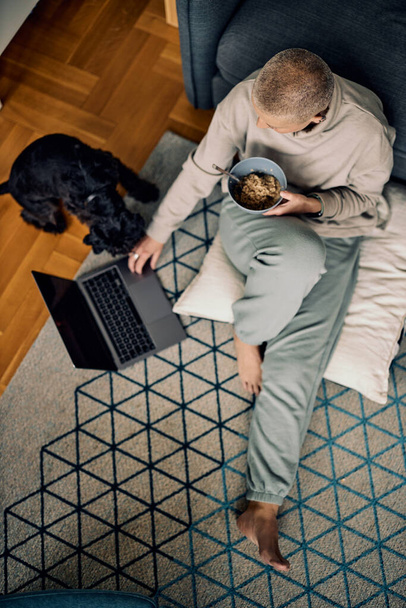 Draufsicht einer erwachsenen Frau mit kurzen Haaren, die einen Laptop benutzt, während sie auf dem Boden sitzt und frühstückt. Neben ihr versucht ihr geliebter Hund, mit ihr zu spielen. - Foto, Bild