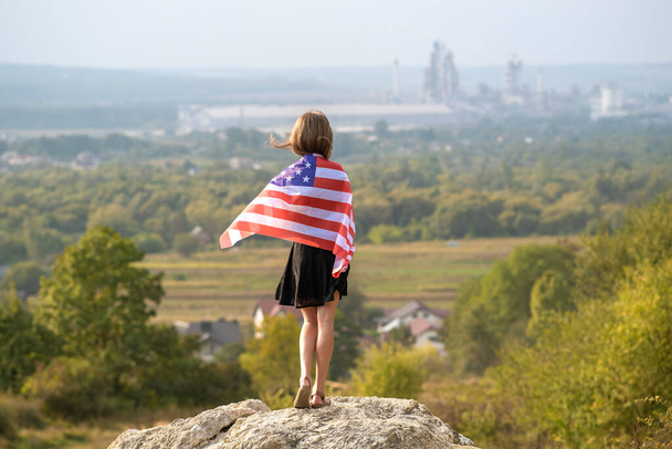 Junge hübsche Amerikanerin mit langen Haaren und wehender US-Flagge auf ihren Schirmen, die draußen steht und den warmen Sommertag genießt. - Foto, Bild