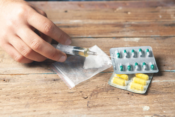 наркоман держит шприц с жидкостью, а на столе пакет с порошком и таблетками. Концепция дополнения - Фото, изображение
