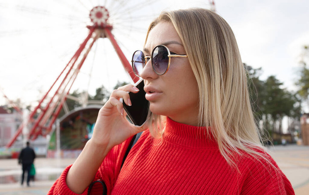 attractive sexy blonde girl in red top and sunglasses using smartphone in amusement park  - Фото, зображення