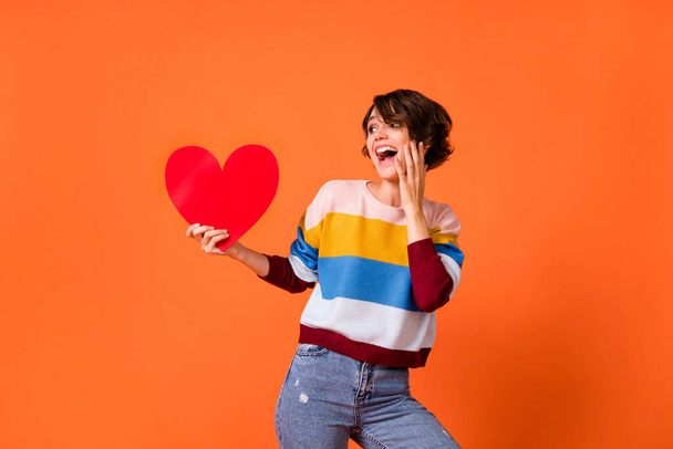 Photo of excited adorable girl hold paper heart paper look empty space hand on cheek isolated on orange color background - 写真・画像