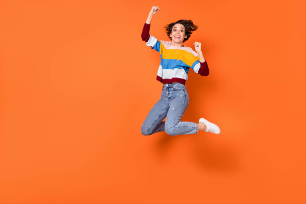 Full length photo of delighted girl fists up celebrate jumping high isolated on orange color background - Photo, image