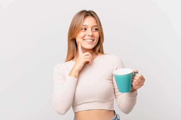 young woman with a coffee mug smiling with a happy, confident expression with hand on chin - Photo, image