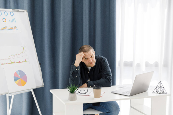 A man dressed in casual clothes works in the office with a laptop and expresses sad emotions. The man works in a remote place of work - Photo, Image