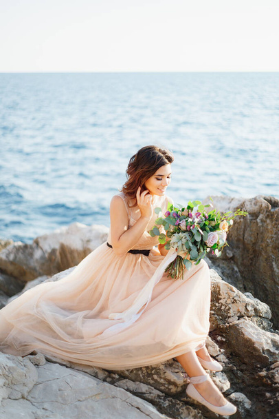 Mooie glimlachende bruid in een beige jurk met een boeket bloemen in haar handen zit op een rots boven de zee - Foto, afbeelding