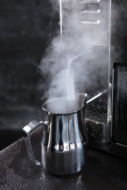 A metal milk jug next to the coffee machine. A strong jet of steam. Vertical photo. - Fotó, kép