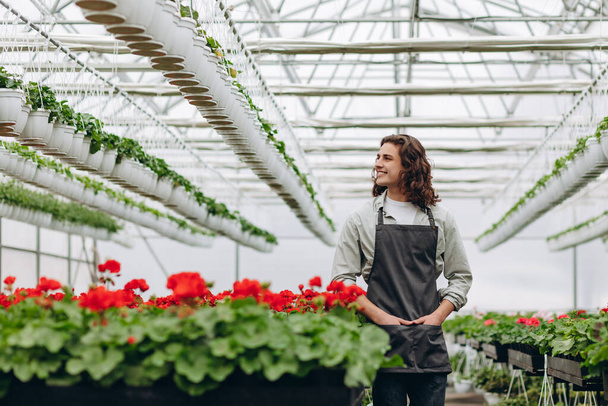Счастливый рабочий, в фартуке, выращивающий цветы в оранжерее цветочного магазина - Фото, изображение