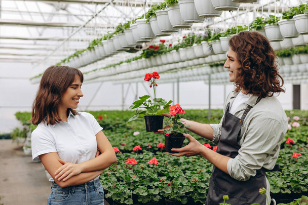 Imagen de un hombre joven parado en el invernadero con una mujer sosteniendo plantas. Mirando a un lado - Foto, imagen