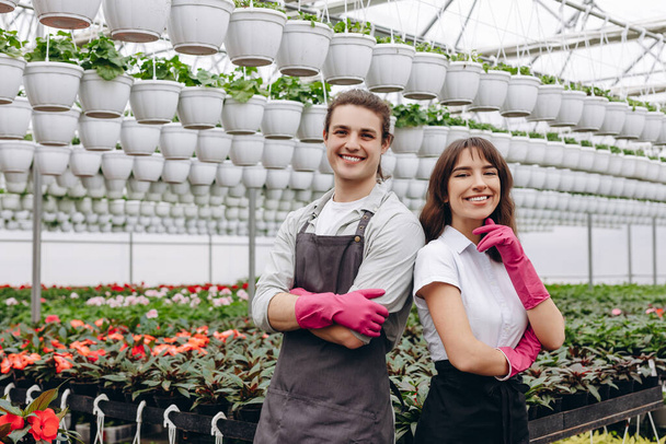 Mannelijke en vrouwelijke bloemisten, met schort en handschoenen aan, glimlachend en kijkend naar de camera, werkend in kas, tuincentrum - Foto, afbeelding