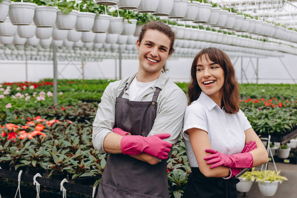 Férfi és női virágárusok kötényben és kesztyűben, mosolyogva és kamerába nézve, miközben üvegházban, kertészeti központban dolgoznak - Fotó, kép