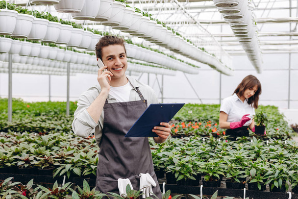 Attractive male florist talking on the phone and holding clipboard, while another female florist arranging pots with flowers on background in greenhouse - Valokuva, kuva