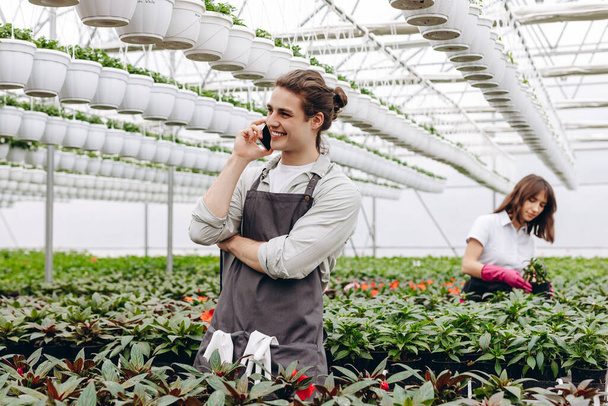 Atractiva florista masculina hablando por teléfono y sujetando el portapapeles, mientras que otra florista femenina arreglando macetas con flores en el fondo en invernadero - Foto, Imagen