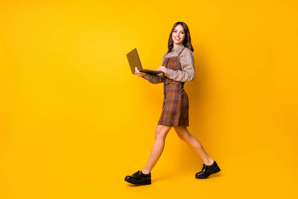Full size profile photo of optimistic brunette lady go laptop wear brown shirt dress boots isolated on vivid yellow background - Photo, image