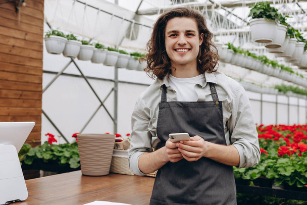 Jonge glimlachende bloemist glimlachen en het gebruik van de smartphone tijdens het werken in de bloemenwinkel - Foto, afbeelding