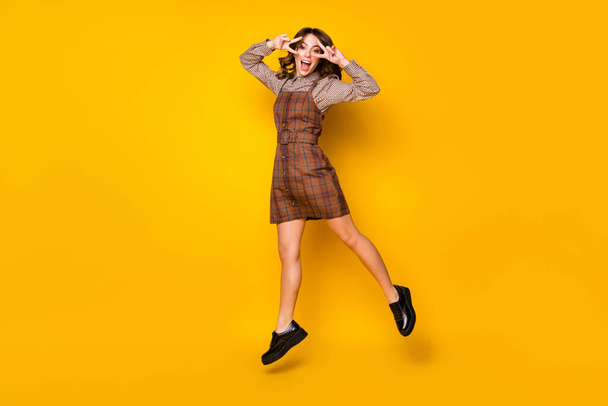 Full size photo of optimistic funny brunette lady jump show v-sign wear shirt dress sneakers isolated on vivid yellow background - Foto, Imagem