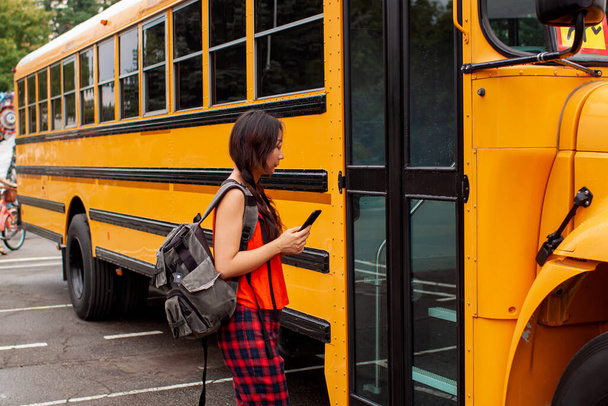 Ασιάτισσα έφηβη στέκεται δίπλα σε μια μεγάλη κίτρινη πόρτα σχολικού λεωφορείου με την τσάντα της. - Φωτογραφία, εικόνα