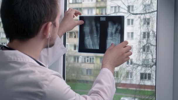 Junger Arzt beim Betrachten eines Röntgenbildes, vor dem Hintergrund eines Fensters, vor dem Hintergrund eines Gebäudes, des Therapiekonzepts und eines Arztes - Filmmaterial, Video