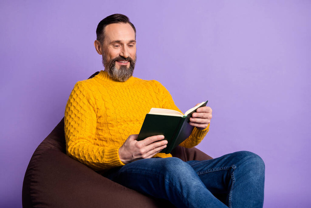 Portrait of cheerful man sitting soft chair hands hold read book toothy smile isolated on purple color background - Foto, afbeelding