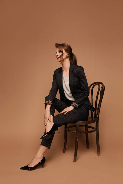 Adult woman, caucasian business lady in a grey suit, blouse and black shoes posing on the chair over beige background, isolated with copy space - 写真・画像