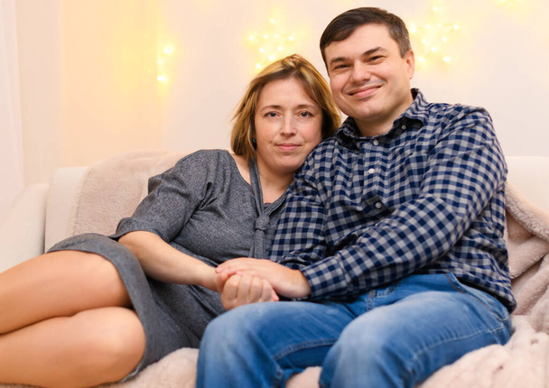 portrait of adult romantic couple sitting on a couch at home, holiday lights on a wall - Foto, imagen