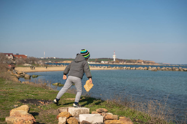 Ένα πεντάχρονο αγόρι πετάει πέτρες στη θάλασσα. Ψυχαγωγία κατά την απομόνωση - Φωτογραφία, εικόνα