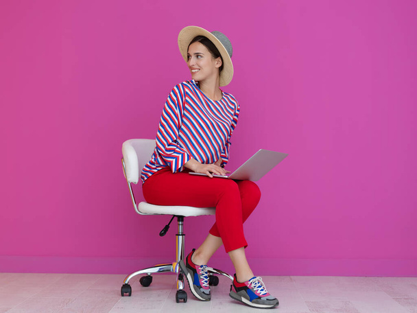 Portrait of young woman sitting on the chair and holding laptop on the lap isolated on pink background. Female model presenting fashion and technologz concept - Photo, image