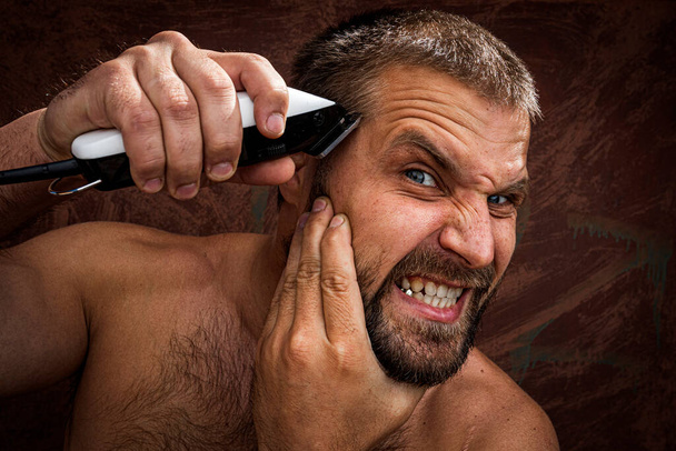 Ritratto ravvicinato di un bell'uomo senza maglietta che si depila la testa con un rasoio elettrico e stringe i denti, su uno sfondo brutale. concetto di assistenza domiciliare maschile senza saloni di bellezza - Foto, immagini