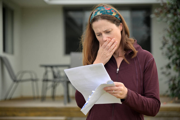 An upset adult woman (female age 25-35) reading a letter in home front yard. - Photo, Image