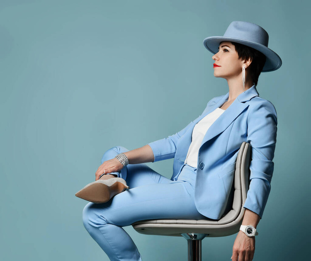 Smiling brunette woman in blue business suit, hat and high-heeled shoes sits relaxed on chair looking at copy space - Foto, Imagem