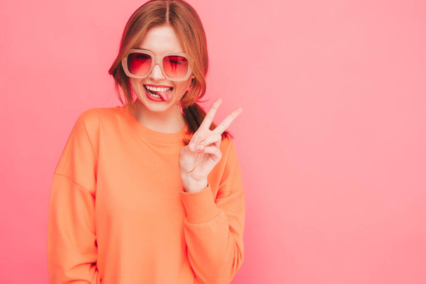 Young beautiful smiling female in trendy summer clothes. Sexy carefree woman posing near pink wall in studio. Positive model having fun in sunglasses. Cheerful and happy.Shows tongue and peace sign - Foto, imagen