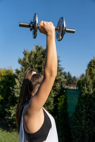 A sporty woman holding a dumbbell and working out in the park - Foto, imagen