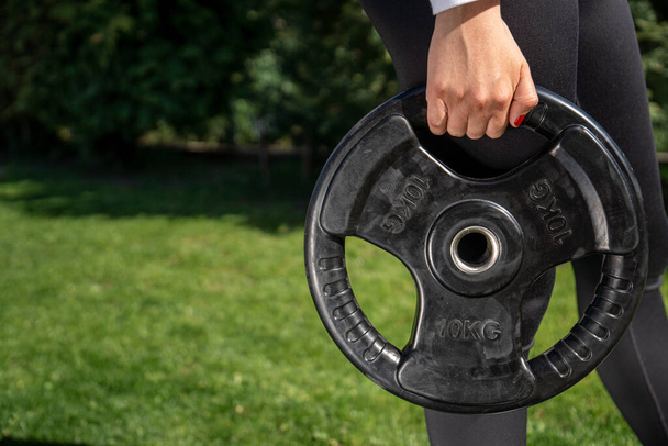An unrecognizable woman holding a 10 kg barbell disc with handles and working out in the park - 写真・画像