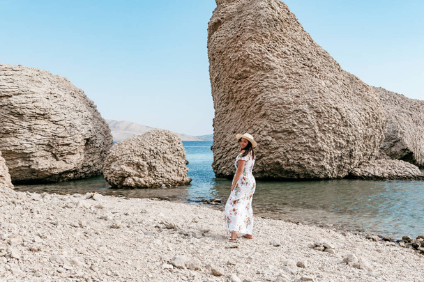 Kobieta w eleganckiej długiej białej sukience letniej stojąca na pięknej plaży Beritnica z dużymi formacjami skalnymi na wyspie Pag w Chorwacji. - Zdjęcie, obraz