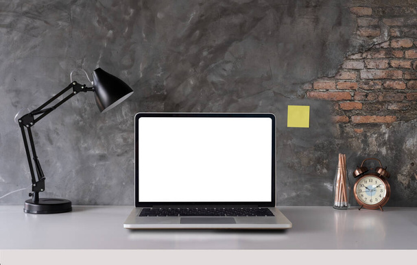 Escritorio blanco con computadora portátil pantalla blanca en blanco, lámpara, suministros de oficina, reloj en jarrón de diseño - Foto, Imagen